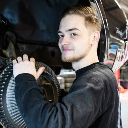 Ein junger Mann lächelt, während er an einem Autoreifen arbeitet; er trägt einen schwarzen Pullover.