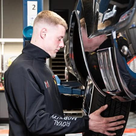 Junger Mann prüft konzentriert einen Autoreifen, trägt schwarze Arbeitsjacke, helles Haar.