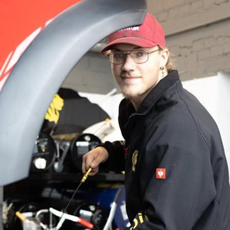 Ein Mann mit Brille und rotem Hut lächelt. Er trägt eine schwarze Jacke und arbeitet an einem Auto.