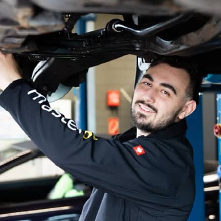 Ein Mechaniker lächelt, während er ein Auto inspiziert. Er trägt eine schwarze Jacke.